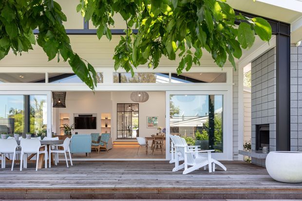 A bright contemporary outdoor entertaining area opening directly into an open-plan living and dining space. The broad timber deck has white dining furniture, lounge chairs and planters, while the house features extensive glazing, pale horizontal cladding, a dark tiled fireplace wall, and a covered upper level. Mature leafy branches frame the view.