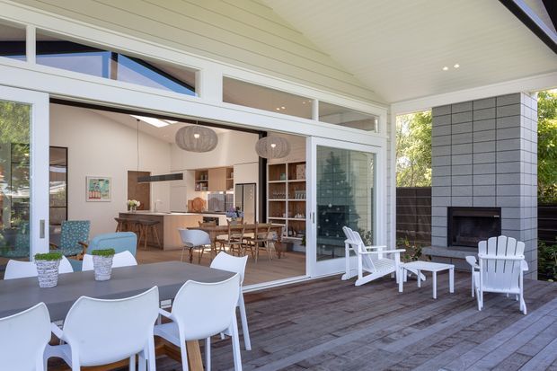 Bright covered outdoor entertaining deck opening through large sliding glass doors into an open-plan kitchen and dining area. The deck features a dining table, white outdoor lounge chairs, a built-in dark fireplace set into a pale masonry wall, and timber decking. The interior has light cabinetry, timber accents, pendant lights, and extensive glazing beneath a white-lined ceiling.