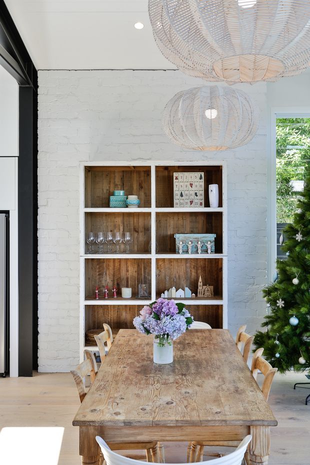 Bright dining area with a long rustic timber dining table and matching wooden chairs, set in front of built-in open shelving with white framing and warm timber backing. The room features a white-painted exposed brick wall, black-framed glazing or doors at left, pale timber flooring, and large woven pendant lights. A decorated Christmas tree and floral centerpiece add seasonal accents.