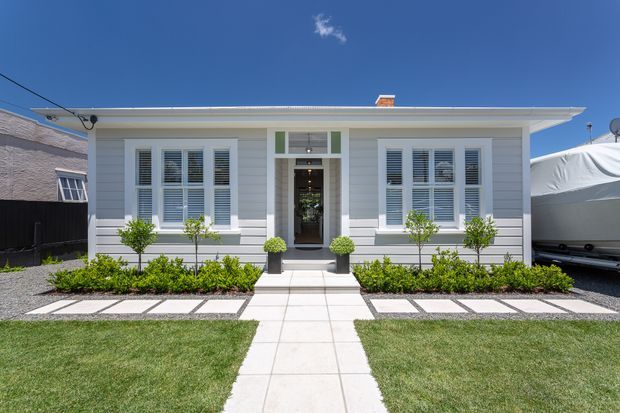Front exterior view of a compact, single-storey light-grey weatherboard house with a low-pitched roof, symmetrical facade, white-framed shuttered windows, recessed central entry, and a formal landscaped front garden. A straight tiled path leads through lawns and gravel garden beds to the entrance.