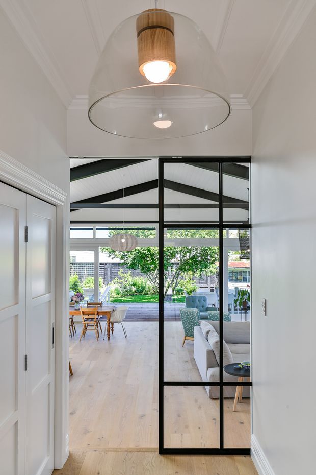 View from a hallway or entry passage through a black-framed glazed partition into a bright open-plan living and dining area. The interior features white walls and ornate ceiling cornices, pale timber flooring, a modern glass pendant with timber detail, contemporary sofas and armchairs, and a dining table beyond. Large glazing opens toward a landscaped garden and patio.