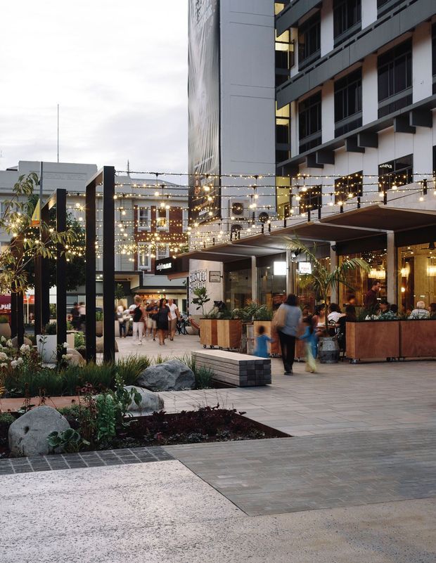 An urban mixed-use outdoor plaza at dusk, framed by a contemporary multi-storey building with balconies and dark horizontal detailing. The pedestrian space includes paved terraces, raised planters, ornamental grasses, palms, rocks, benches, restaurant frontage, and extensive warm string lighting. Outdoor dining areas and blurred pedestrians create an active, inviting hospitality atmosphere.