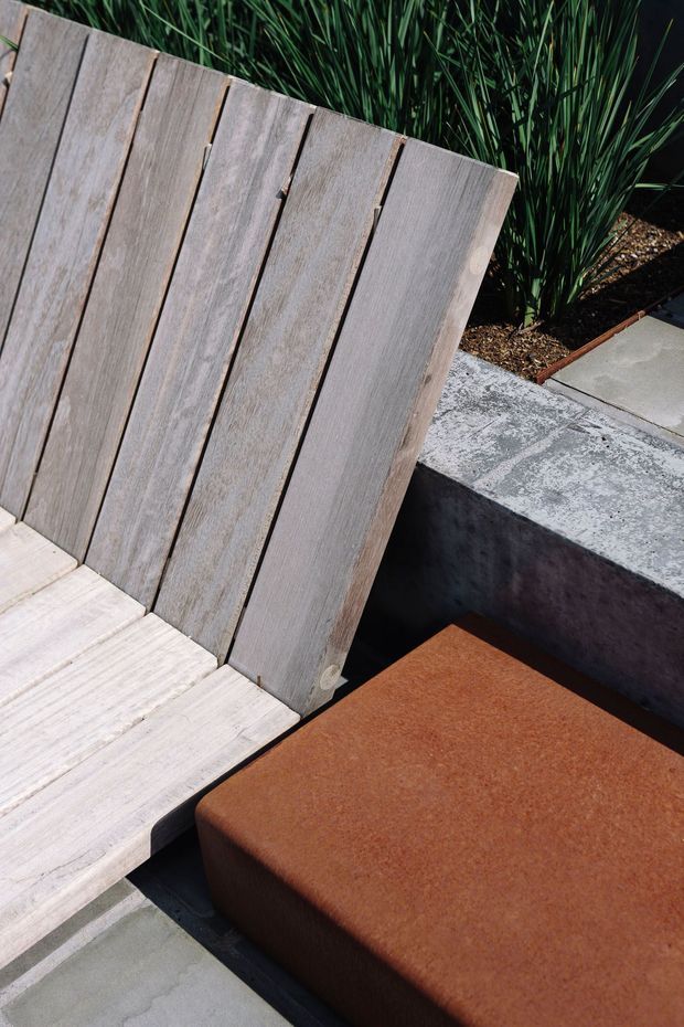 Close-up outdoor seating area featuring a weathered light timber slatted lounge chair, a rust-brown upholstered ottoman or cushion, and a dark concrete bench edge. Grey concrete paving, raised garden planting, and a restrained natural material palette create a contemporary landscaped patio setting.