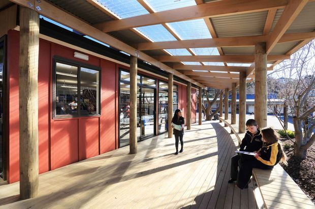 A covered outdoor walkway beside a contemporary low-rise building, featuring red wall panels, extensive glazing, exposed timber posts and beams, and translucent roof panels that provide filtered daylight. Timber decking curves around built-in bench seating, with people occupying the sheltered circulation space. The scene has a warm, functional institutional or commercial character.