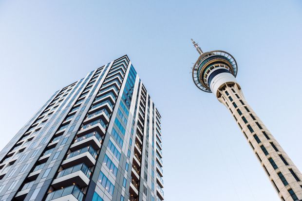 Low-angle exterior architectural photograph showing a tall modern high-rise building with a glass-and-panel facade and projecting balconies beside a distinctive telecommunications observation tower. The scene is dominated by contemporary urban vertical architecture against a clear blue sky.