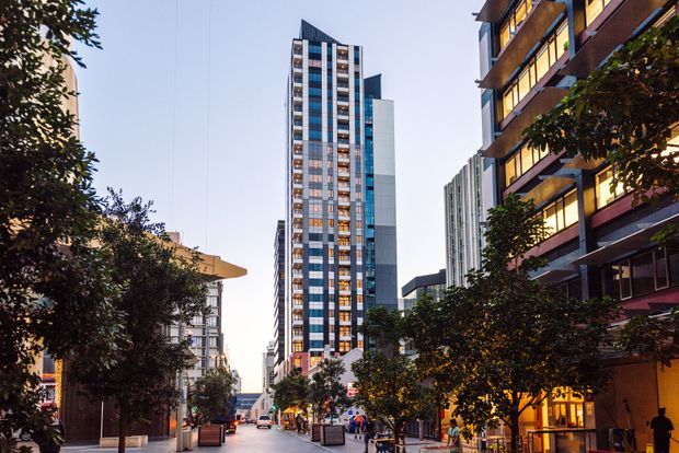 Urban streetscape viewed along a pedestrian-oriented city street, framed by mature trees and lined with contemporary mid- and high-rise apartment or commercial buildings. A tall narrow tower with extensive vertical glazing and patterned facade panels dominates the centre, while illuminated balconies and shopfront-like spaces appear on the right. The scene has a lively, polished metropolitan character in late-day light.