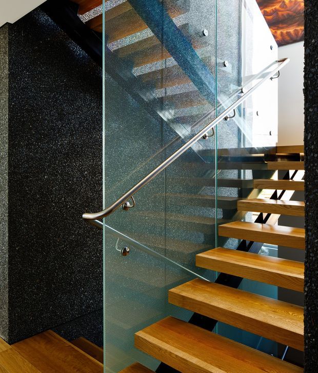 Interior view of a modern open-riser staircase with thick timber treads, a clear glass balustrade, and a polished metal handrail. Dark textured feature walls contrast with the warm timber and transparent glazing. The stair structure and upper landing are visible through the glass, creating a sculptural, contemporary architectural composition.