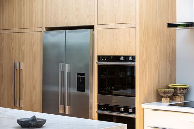 Modern kitchen with full-height light timber cabinetry, integrated stainless-steel French-door refrigerator, stacked black-glass ovens, and a pale stone island benchtop. The image also shows a white drawer unit with a dark cooktop and decorative woven vessels.