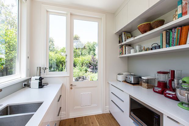 Bright, compact galley-style kitchen with white cabinetry, open shelving, a white benchtop, stainless-steel sink, integrated appliances and colourful countertop appliances. Full-height glazing and a glazed timber door bring in abundant natural light and overlook greenery. Timber flooring adds warmth.