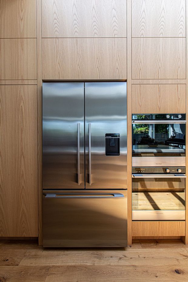 Close-up view of a contemporary kitchen wall featuring full-height natural timber cabinetry, a large stainless-steel French-door refrigerator, and integrated stainless-steel ovens. Warm timber flooring and strong sunlight create a refined, modern kitchen interior.