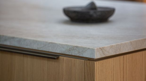 Close-up detail of a contemporary built-in cabinet or kitchen island, featuring a pale natural-stone benchtop with a thick edge, warm vertical-grain timber cabinetry, and a slim dark recessed handle. A shallow dark decorative bowl sits on the benchtop.