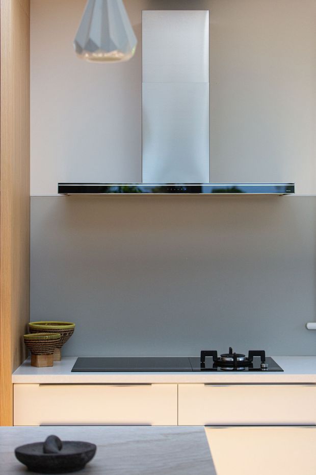 A close-up view of a contemporary kitchen cooktop set into pale cabinetry, with a sleek black gas and induction-style hob, integrated stainless-steel rangehood, smooth grey splashback, light timber side panel, and a faceted pendant light. Woven bowls and a dark decorative bowl add subtle natural accents.