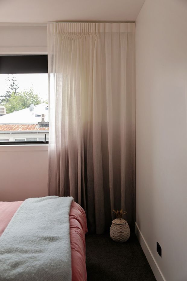 A compact, softly styled bedroom corner with a partially visible bed dressed in blush pink bedding and a pale blue textured throw. Floor-to-ceiling sheer curtains in a taupe-to-charcoal gradient frame a window, with dark carpet underfoot, white walls and ceiling, and a small woven decorative planter beside the curtains. The room has a warm, muted, intimate feel.