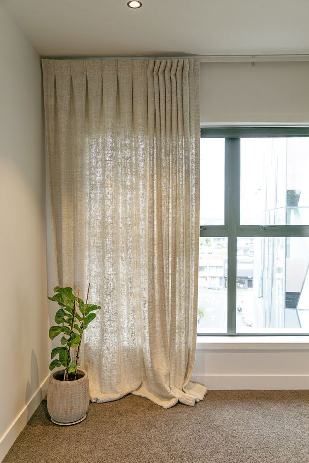 A softly lit, neutral-toned interior corner featuring full-height textured beige curtains beside a large grey-framed window. A potted indoor plant sits on beige carpet, with plain painted walls, a recessed ceiling light, and simple contemporary detailing.