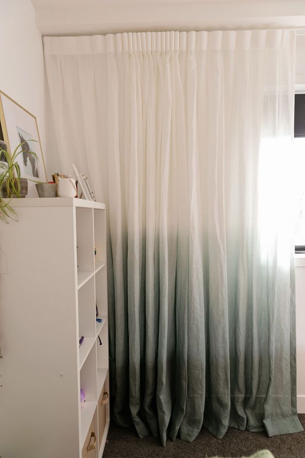 A bright, softly styled interior corner featuring full-height pleated curtains with a dramatic white-to-deep green gradient. A white open shelving unit with small objects and a potted plant sits to the left, while daylight enters through a window on the right. The room has white walls and dark neutral carpet, creating a calm, contemporary atmosphere.
