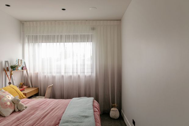 A compact, softly lit bedroom with a single bed dressed in pink bedding and a pale blue throw, sheer full-height curtains over a large window, and a small timber desk or shelf with decorative items. The room has plain light walls, recessed ceiling lights, carpeted flooring, and a calm pastel palette.
