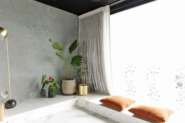Bright contemporary interior corner featuring a built-in white marble-look window seat beneath floor-to-ceiling glazing. The space has a grey textured concrete-look wall, dark ceiling, sheer full-height curtains, potted plants, and rust-orange cushions. Decorative patterned glazing creates soft filtered light.