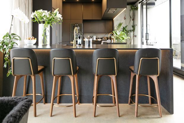 Contemporary open-plan kitchen dominated by a large dark island with four upholstered bar stools. The space features dark flat-panel cabinetry, integrated appliances, pendant-free recessed lighting, a pale stone or concrete-look floor, extensive glazing, and abundant indoor greenery and floral arrangements.