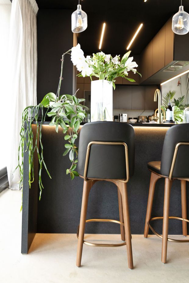 A stylish compact kitchen and breakfast bar with a dark matte island, integrated cabinetry, black ceiling, warm timber accents, and brass-toned fixtures. Two upholstered timber bar stools sit at the island, while pendant lights and linear task lighting illuminate the space. Indoor plants and a large floral arrangement add a natural decorative element beside full-height sheer curtains.