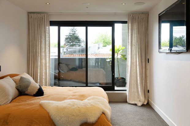 A warm, contemporary bedroom with a low-profile bed dressed in ochre bedding and layered cream sheepskin throws. Full-height black-framed glazed doors open to a small balcony or terrace, with textured floor-length curtains, neutral carpet, recessed ceiling lights, and a wall-mounted television. The room receives abundant natural light and overlooks an urban residential setting.