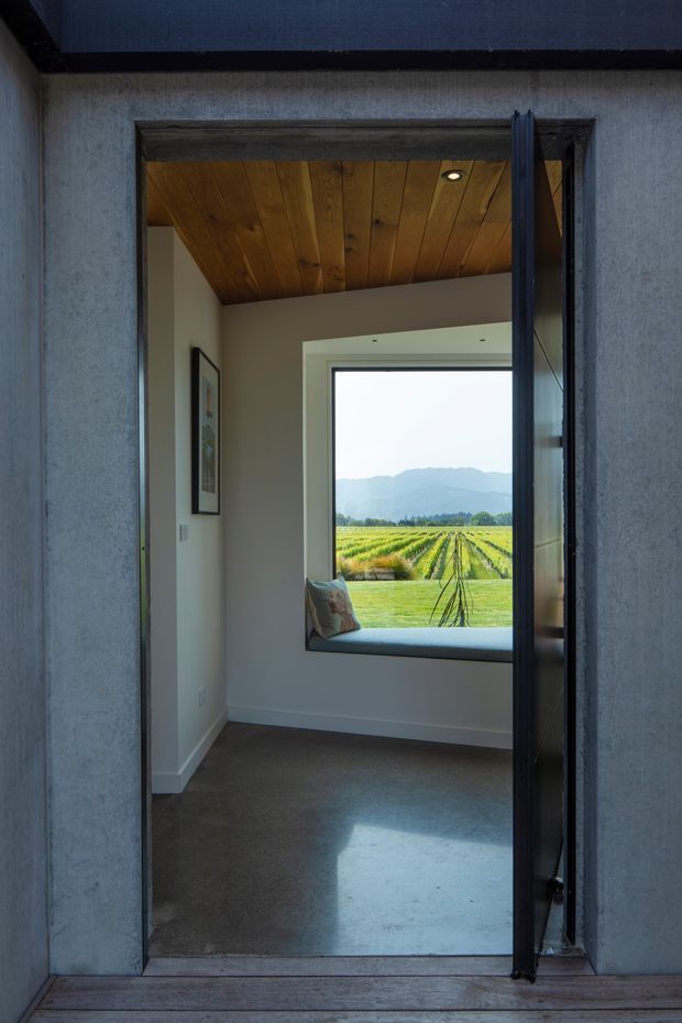 View through an open dark-framed doorway into a compact, quiet interior space with a grey carpeted floor, white walls, a timber-lined sloping ceiling, and a built-in window seat. The large window frames rows of cultivated fields and distant mountains. A timber deck and textured grey exterior wall are visible around the doorway.