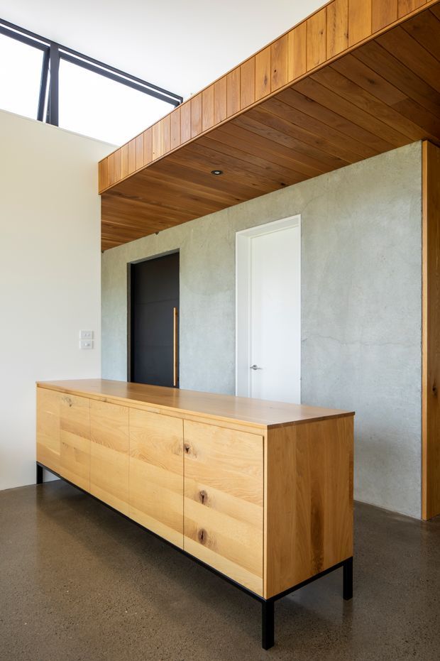 A refined interior entry or hallway featuring a large light timber sideboard on black legs, a textured grey plaster wall, contrasting black and white doors, and a dramatic timber-lined ceiling and overhead built-in joinery. High clerestory glazing brings in abundant natural light. The palette is warm timber, concrete grey, black, and white, with a clean mid-century-inspired architectural character.