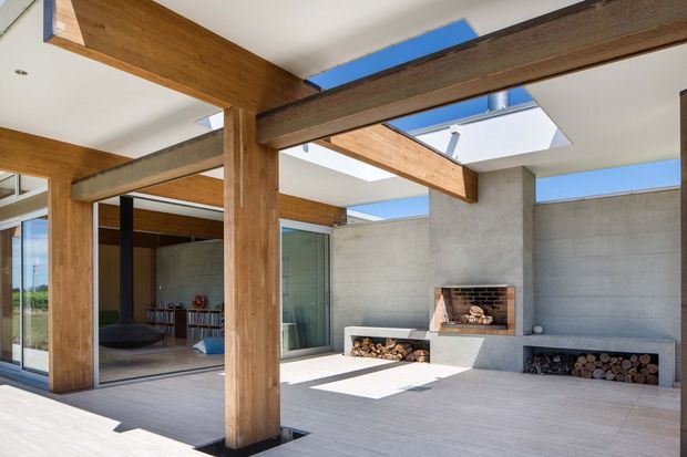Bright contemporary indoor-outdoor living space with exposed timber posts and beams, smooth concrete walls, large glazed openings, skylights, and a built-in masonry fireplace with integrated firewood storage. The open terrace-like area is sunlit and visually connected to an interior lounge through full-height glazing.