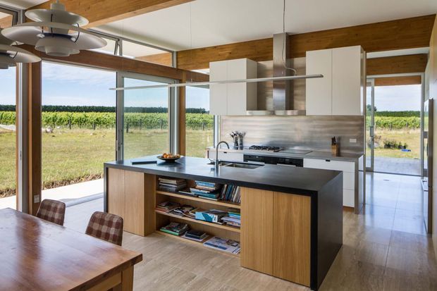 A bright open-plan kitchen and dining area with a large central island, integrated sink, white overhead cabinetry, stainless-steel splashback and appliances, and warm timber cabinetry. Full-height glazing opens the room to a rural vineyard landscape, while exposed timber ceiling beams and a contemporary pendant light add architectural and decorative interest.