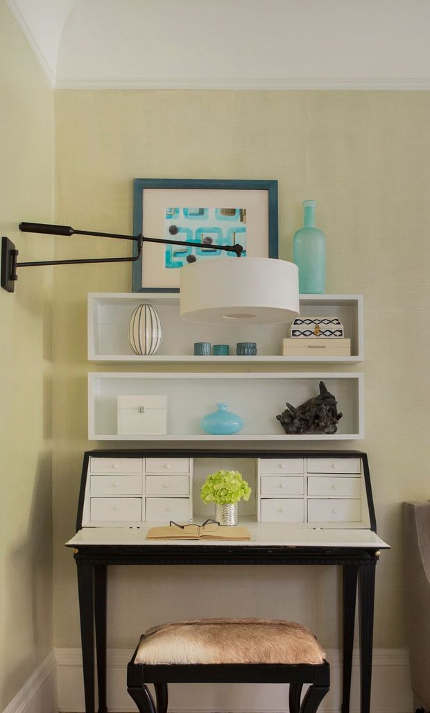 A compact, traditionally detailed writing and dressing nook against a pale green wall. A dark wood console desk with white built-in drawers sits beneath two white open display shelves, styled with books, ceramics, a vase, artwork and a sculptural object. A black swing-arm wall lamp and large white drum pendant provide lighting, while a cushioned dark stool is tucked beneath the desk. Decorative crown and base mouldings frame the space.
