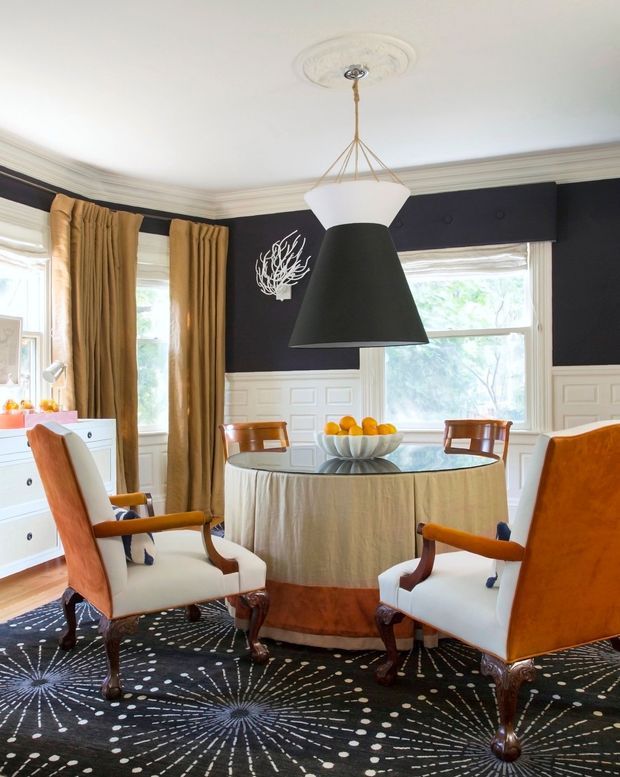 Elegant formal dining room with a round glass-topped dining table draped in beige fabric, surrounded by upholstered wood-framed armchairs. The room features dark charcoal walls, white decorative wall panelling and ornate ceiling cornices, gold curtains, a dramatic black-and-white pendant light, and a bold patterned dark rug.