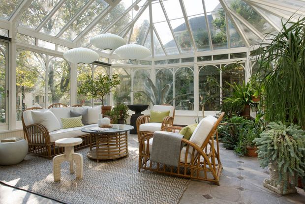 A bright, glass-enclosed conservatory or sunroom filled with lush indoor plants. The space features a glazed pitched roof and framed arched windows, pale stone flooring, a patterned area rug, and a relaxed seating arrangement of woven rattan and upholstered armchairs around a low coffee table. Sculptural pendant lights hang overhead, creating an airy indoor-outdoor atmosphere.