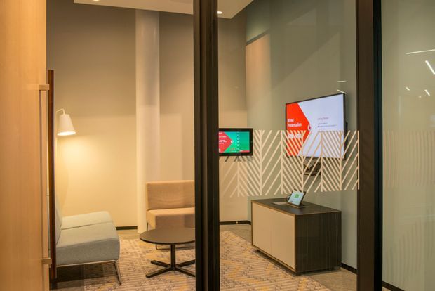 A modern commercial reception or waiting area viewed through full-height glazed doors or partitions. The space includes upholstered lounge seating, a floor lamp, a compact reception counter with a tablet, digital screens, and a decorative geometric wall pattern. Warm ambient lighting, dark framing, and patterned carpet create a refined contemporary office atmosphere.