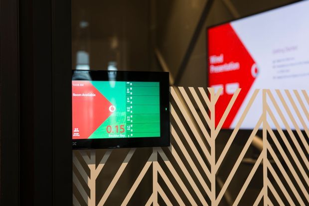 A contemporary commercial interior showing a digital room-availability display mounted on a dark wall, with bold geometric black-and-white decorative graphics and a red-and-green interface on the screens. The space appears to be a meeting-room or workplace wayfinding area.