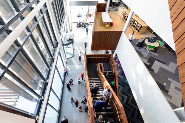 High-angle view of a bright multi-level commercial interior, likely an office or institutional atrium. Floor-to-ceiling glazing lines one side, with an open circulation space, sculptural timber-lined stair, elevated timber-clad platforms, workstations, meeting areas, and patterned carpet. People are present throughout the space.