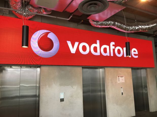 Interior view of a commercial Vodafone-branded lift lobby, with multiple stainless-steel elevator doors set into a raw concrete wall. A large illuminated red Vodafone sign dominates the upper wall, while the exposed ceiling reveals ductwork, services, and suspended black cylindrical pendant lights.