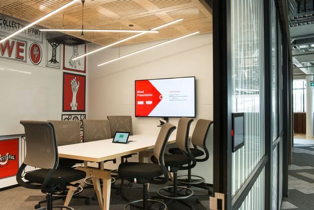 A modern commercial meeting room with a long white conference table, multiple ergonomic office chairs, a wall-mounted presentation screen, and sports-themed wall graphics. The room has a warm slatted timber acoustic ceiling with integrated linear lighting, glass partition walls with vertical privacy fins, and a carpeted floor.
