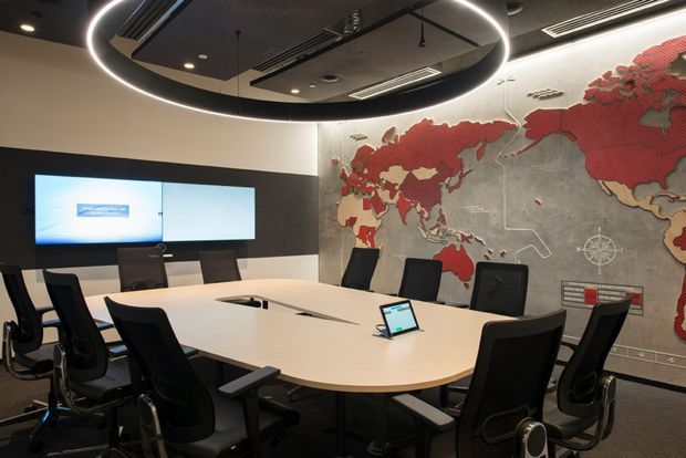 Contemporary corporate boardroom with a large oval white conference table surrounded by black ergonomic office chairs. A wall-mounted display sits against a dark feature wall, while a large textured world map mural creates a prominent feature wall. The room has a dark exposed-services ceiling, recessed and linear lighting, and a large circular ring pendant light.