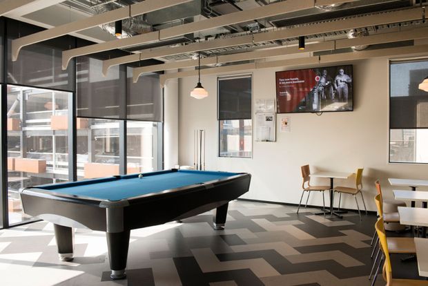 A bright contemporary commercial recreation and breakout space with a large pool table, café-style tables and chairs, expansive windows, exposed services, suspended linear lighting, and geometric patterned carpet tiles. A wall-mounted digital display and glazed partitions contribute to the office or institutional setting.
