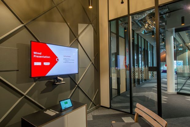A contemporary commercial presentation or meeting area with a wall-mounted display showing a wired presentation, set against a dark geometric wall panel. A small presentation desk with laptop and conferencing equipment sits below. Glazed black-framed meeting-room partitions open onto a modern office corridor, with exposed services and patterned carpet visible beyond.