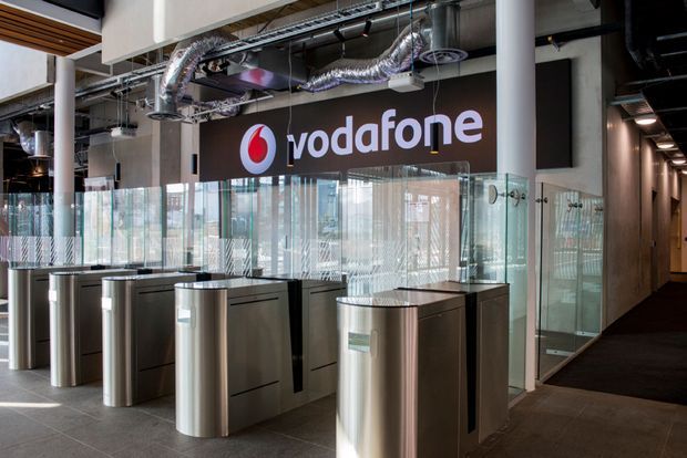 A modern Vodafone office reception and access-control area with stainless-steel security turnstiles, glass partitions, a branded feature wall, exposed mechanical services, and a corridor extending to the right. The space has an industrial commercial fit-out with a neutral palette and polished metal finishes.
