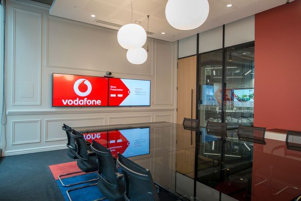 A contemporary commercial meeting or presentation room featuring a long glossy dark table, black mesh conference chairs, wall-mounted Vodafone presentation screens, decorative wall panelling, black-framed glass partitions, and large white spherical pendant lights. The palette combines pale grey wall mouldings with bold red branding and reflective dark surfaces.