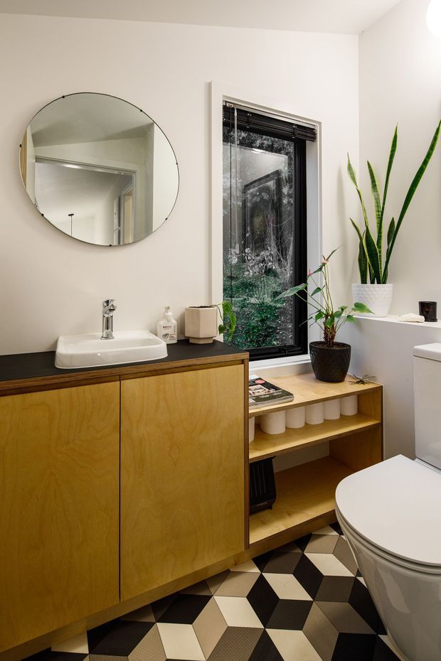 A stylish compact bathroom with a floating timber vanity, black benchtop, white vessel sink and chrome tapware. A large round mirror sits above the vanity, while a tall black-framed window brings in views of greenery. Open timber shelving provides toilet-paper storage beside a contemporary toilet. The floor features a bold geometric pattern in black, white and beige, complemented by potted plants and warm neutral walls.