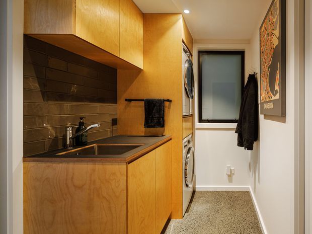 Compact, warm-toned laundry room with integrated timber cabinetry, a dark tiled splashback, sink and benchtop on the left, and stacked front-loading washer and dryer in a central cabinet. A narrow corridor leads to a frosted window, with wall hooks, hanging garments and framed artwork.