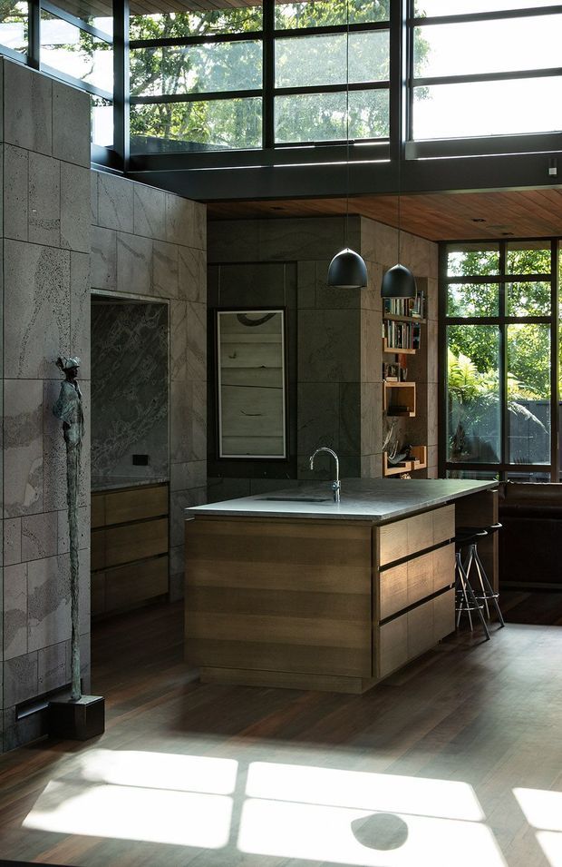 A sophisticated open-plan kitchen with a central timber island, dark stone benchtop, integrated sink and brushed metal tap. The room features extensive black-framed glazing, a warm timber ceiling, dark timber flooring, large-format grey stone wall cladding, pendant lights, open shelving and a sculptural decorative figure. Strong natural light filters through the surrounding trees, creating a dramatic indoor-outdoor connection.