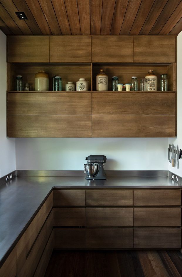 A compact, highly resolved kitchen with dark timber cabinetry wrapping the walls and forming a U-shaped work area. The upper joinery includes open display niches containing jars and crockery, while the lower cabinets provide extensive drawer storage. A dark stainless-steel or metal-look benchtop runs around the kitchen, with a stand mixer on the rear counter and a wall-mounted knife rack at right. The ceiling is clad in matching timber boards, creating a warm, enveloping appearance. Minimal hardware and integrated joinery give the space a clean contemporary feel.