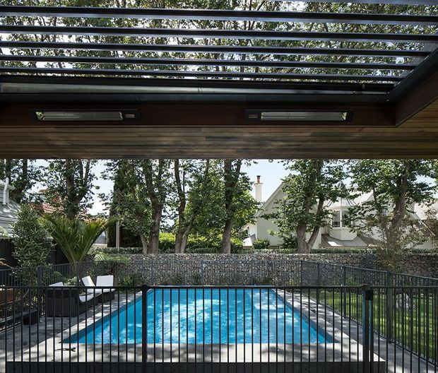 View from beneath a covered outdoor structure with a dark timber-lined ceiling, exposed horizontal roof battens and integrated strip heaters, looking over a rectangular swimming pool enclosed by black metal fencing. Mature trees, a stone boundary wall and neighbouring residential buildings form the background.