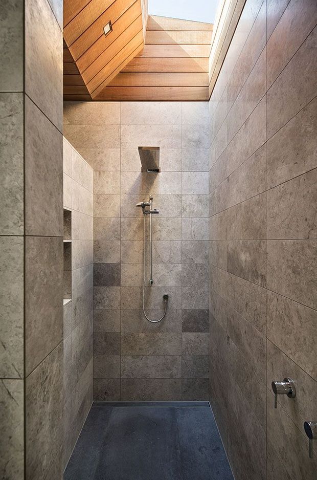 A narrow, minimalist walk-in shower room clad in large-format grey stone-look tiles. The shower features a wall-mounted chrome fixture, recessed wall niches, and a dark tiled floor. A warm timber-lined ceiling with a skylight or clerestory opening brings natural light into the space.