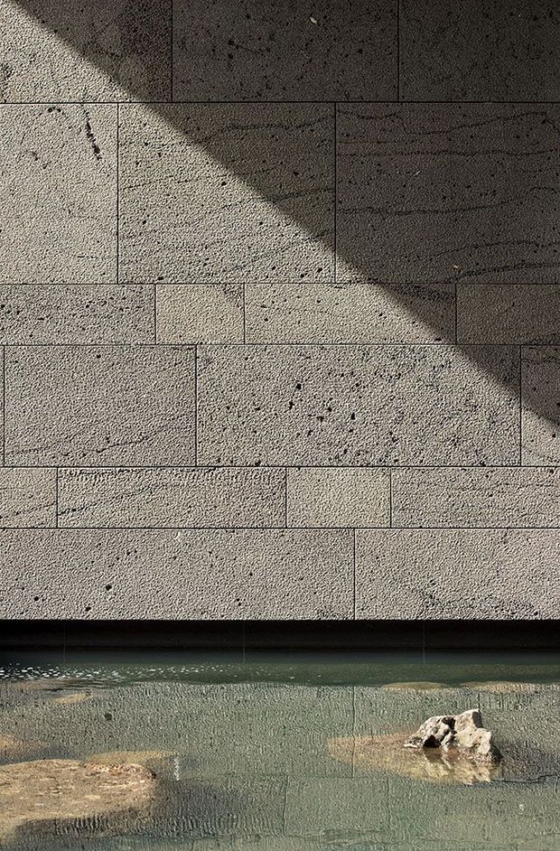 Close-up architectural detail of a textured rectangular stone or concrete block wall above a shallow body of water, with strong diagonal sunlight and shadow creating a dramatic pattern. The water contains partially submerged natural rocks and a dark horizontal wall edge, suggesting a pool or water feature.