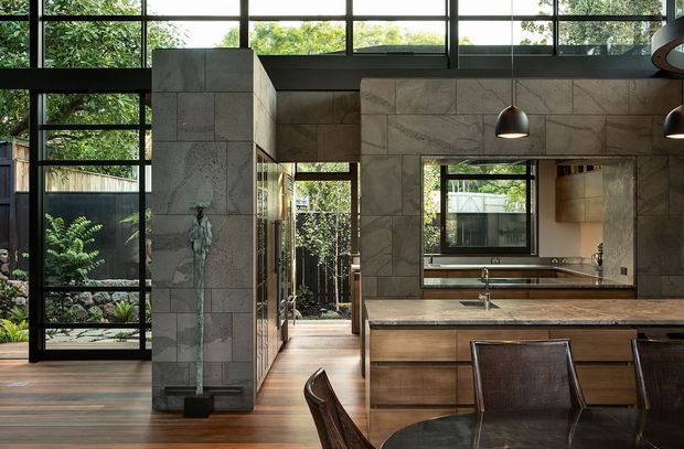 Open-plan contemporary kitchen and dining area with a large timber island, integrated cabinetry, stone-clad feature walls, dark-framed floor-to-ceiling glazing, and pendant lighting. A sculptural vertical stone feature and lush garden are visible beyond the glazed walls, while a mirror reflects additional kitchen joinery.