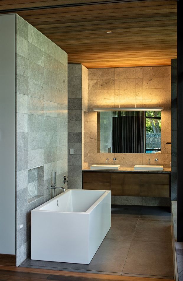 Contemporary bathroom featuring a freestanding white bathtub, double vanity with dark wood cabinetry, large stone-look tiled walls, dark floor tiles, and a warm timber slatted ceiling. A wide mirrored vanity wall reflects greenery and glazing beyond, while concealed and recessed lighting creates a warm, refined atmosphere.